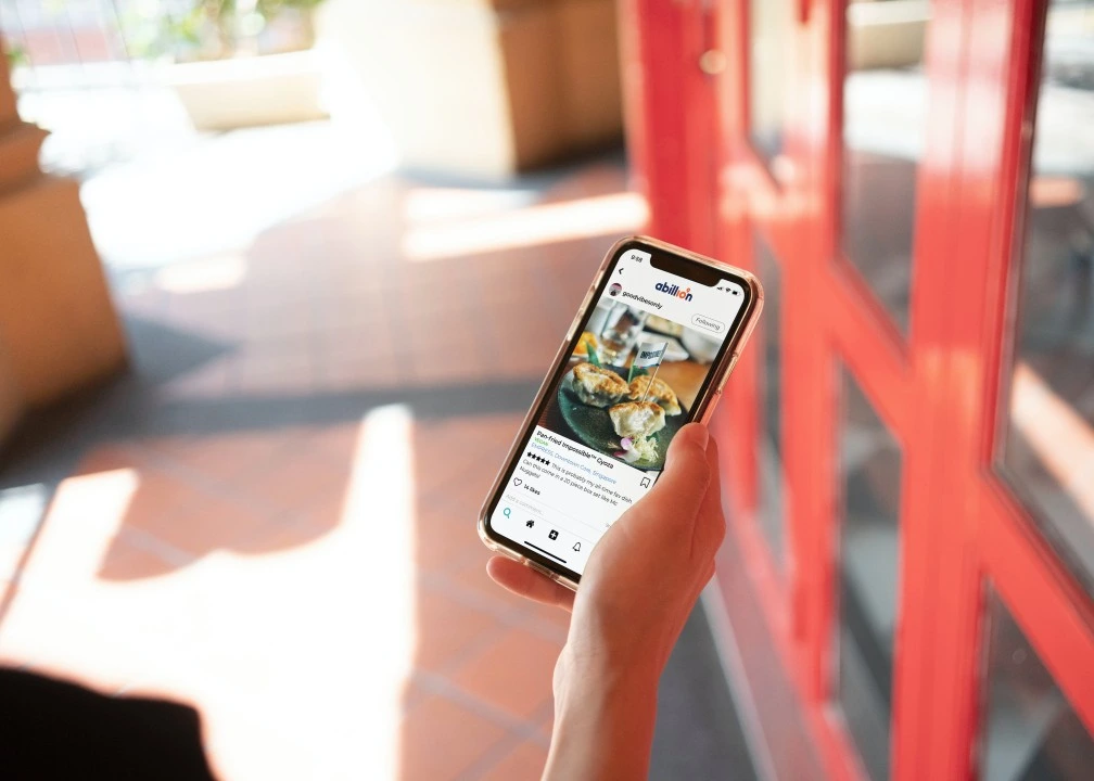 A person holding a smartphone in front of a red building, showing a social media review on the abillion app for a vegan food dish.